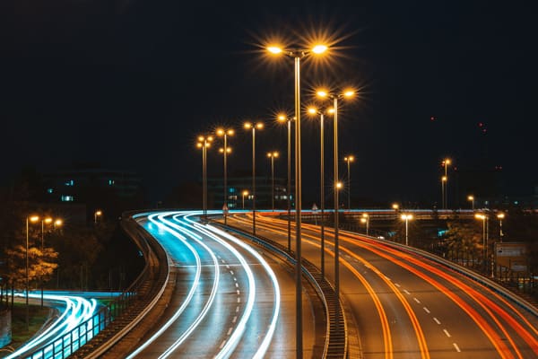 Via urbana iluminada à noite, com rastros de luz e postes acesos